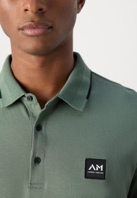 Groene polo met een gestructureerde stof, twee zwarte knopen en een zwart rechthoekig logo met witte letters. Zwarte accenten op de kraag.