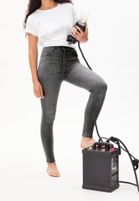 Femme en t-shirt blanc et jean gris tenant un flash de studio avec le pied reposant sur un bloc d’alimentation noir sur fond blanc.