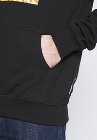 Black sweatshirt with ribbed cuffs and hem, featuring a front pocket. Notable details include vibrant, patterned graphic on the chest.