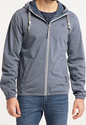 Mand iført grå hættetrøje med lynlås, snore og lommer, kombineret med blå jeans, mod en ensfarvet baggrund.