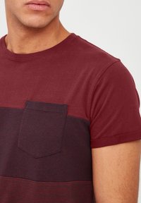 Burgundy t-shirt featuring a color-block design with a front pocket, round neckline, and short sleeves. Soft, smooth fabric texture.