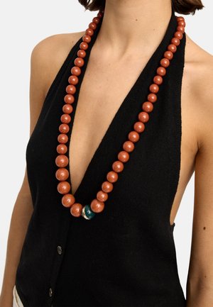 Kobieta ubrana w czarny, bezrękawnik z dekoltem w serek oraz długi naszyjnik z dużych brązowych koralików, z jednym zielonym koralikiem blisko środka.