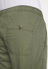 Achterzak met knoop op olijfgroene broek, gedragen met een ingestopte witte shirt, met focus op de stoffen textuur en stikseldetails.