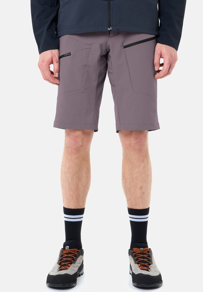 Homme portant un short violet mi-genou avec des poches zippées, des chaussettes noires à rayures blanches, et des chaussures de randonnée grises avec des lacets orange.