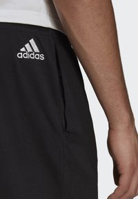 Pantalones cortos deportivos negros con bolsillos laterales, que presentan un logo blanco de Adidas en el muslo izquierdo. La tela parece suave y transpirable.