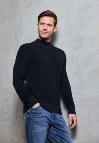 Pull en tricot noir à côtes avec un col montant, associé à un jean en denim bleu. Le tissu semble épais et texturé, se détachant sur un fond gris.