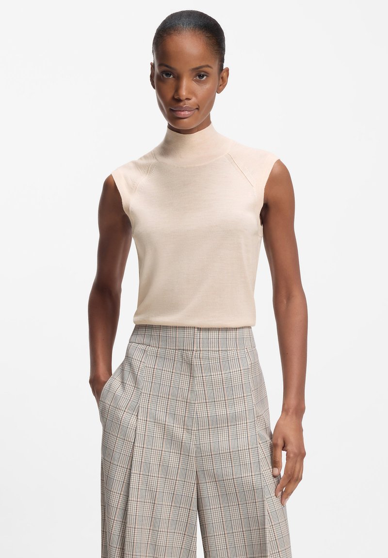 Beżowy bezrękawnik z golfem wykonany z gładkiego materiału, zestawiony z wysokim stanem, spodniami w kratę z delikatnymi czerwonymi akcentami.