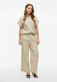 Camicia beige con bottoni e maniche corte, abbinata a pantaloni larghi coordinati. Tessuto liscio con una leggera texture. Scarpe con tacco alto nere.