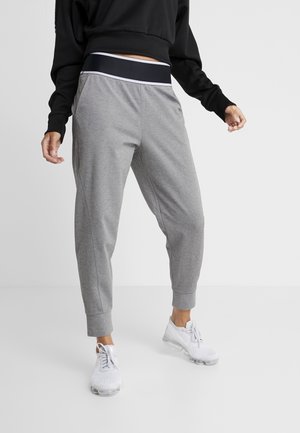 Personne portant un pantalon de survêtement gris avec une ceinture noire et blanche, un haut noir à manches longues et des chaussures de sport blanches, debout devant un fond uni.