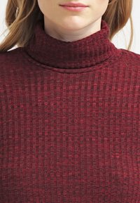 Haut col roulé bordeaux en tricot texturé. Présente un col près du corps et un motif à côtes verticales sur l'ensemble du matériau.