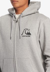 Sudadera con cremallera gris hecha de tela suave, con un logo frontal de una ola y una montaña. Incluye capucha con cordón y bolsillos frontales.