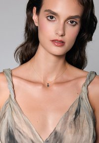 Złoty naszyjnik z niebieskim kamieniem wisiorem. Modelka ma na sobie zwiewną, stonowaną zieloną i beżową sukienkę z delikatnymi fałdami oraz asymetrycznymi ramiączkami.