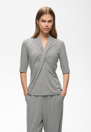 Femme debout, les mains dans les poches, portant un haut drapé gris clair à manches jusqu’aux coudes et un pantalon large assorti, fond neutre.