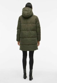 Cappotto trapuntato di colore verde scuro con cappuccio, caratterizzato da pannelli trapuntati e una lunghezza fino al ginocchio. Indossato sopra leggings neri e stivali con tacco.