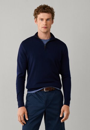 Young man with curly hair wearing a navy blue quarter-zip sweater over a blue shirt and navy pants, standing against a plain background.