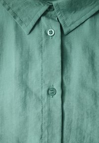 Chemise verte clair à boutons, avec un large col pointu, texture lisse et patte de boutonnage à cinq boutons. Présente un logo rond sur le bouton du haut.