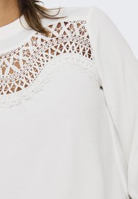 White top featuring intricate lace and cut-out designs along the neckline. Soft texture with long sleeves and a relaxed fit.