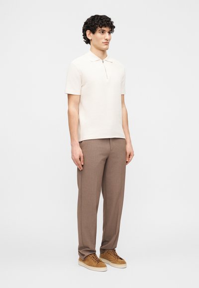 Νεαρός με σγουρά μαλλιά που φοράει ανοιχτό μπεζ κοντομάνικο πόλο με φερμουάρ, καφέ παντελόνι και καφέ σουέντ αθλητικά παπούτσια, όρθιος.