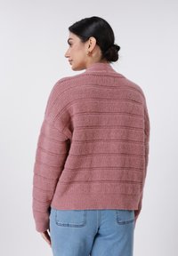 Ροζ πλεκτό πουλόβερ με οριζόντια ρίγα, κομμένο σχέδιο και ψηλό λαιμό. Μαλακή υφή και άνετη εφαρμογή. Φοριέται με ανοιχτόχρωμο μπλε τζιν.