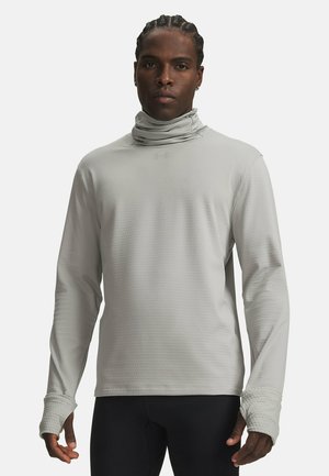 Homme aux cheveux tressés portant un col roulé gris clair à manches longues et un pantalon noir, debout devant un fond uni.