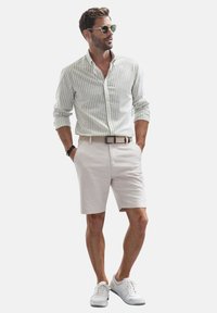 Gestreiftes Herrenhemd in Grün und Weiß, kombiniert mit beigen Shorts. Als Accessoires sind ein Gürtel und eine Sonnenbrille vorhanden. Die Schuhe sind im sportlichen weißen Stil.
