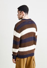 Gestreifter Pullover in Braun, Creme und Marineblau. Strukturiertes Strickmuster mit flauschigen Streifen und gerippten Bündchen. Rundhalsausschnitt.