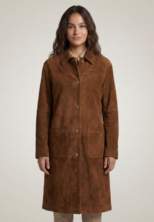 Jeune femme portant un long manteau boutonné en daim marron avec de grandes poches avant, debout devant un fond clair uni.