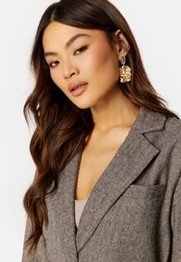 Blazer a zigzag marrone con colletto a revers, caratterizzato da una tasca discreta. Orecchini dorati chunky aggiungono un dettaglio di texture. Capelli morbidi e fluenti.