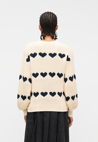 Άτομο  με πλεξούδες, που φοράει κρεμ πουλόβερ με μαύρα σχέδια καρδιάς και σκουρόχρωμη γκριπ φούστα, εμφανίζεται από πίσω με λευκό φόντο.