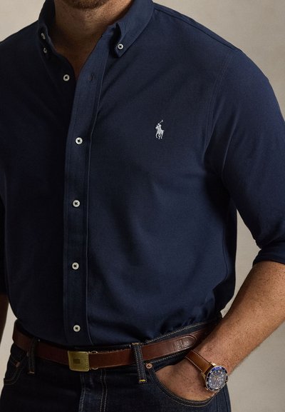 Muškarac u mornarskoplavoj košulji s bijelim logom igrača polo, smeđim kožnim remenom, tamnim trapericama i smeđim satom s plavim brojčanikom, ruka u džepu.