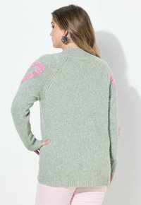 Kobieta stojąca tyłem, ubrana w jasnoszary sweter z dzianiny z różowymi wzorami na rękawach i jasnoróżowe spodnie, ręka oparta na biodrze.