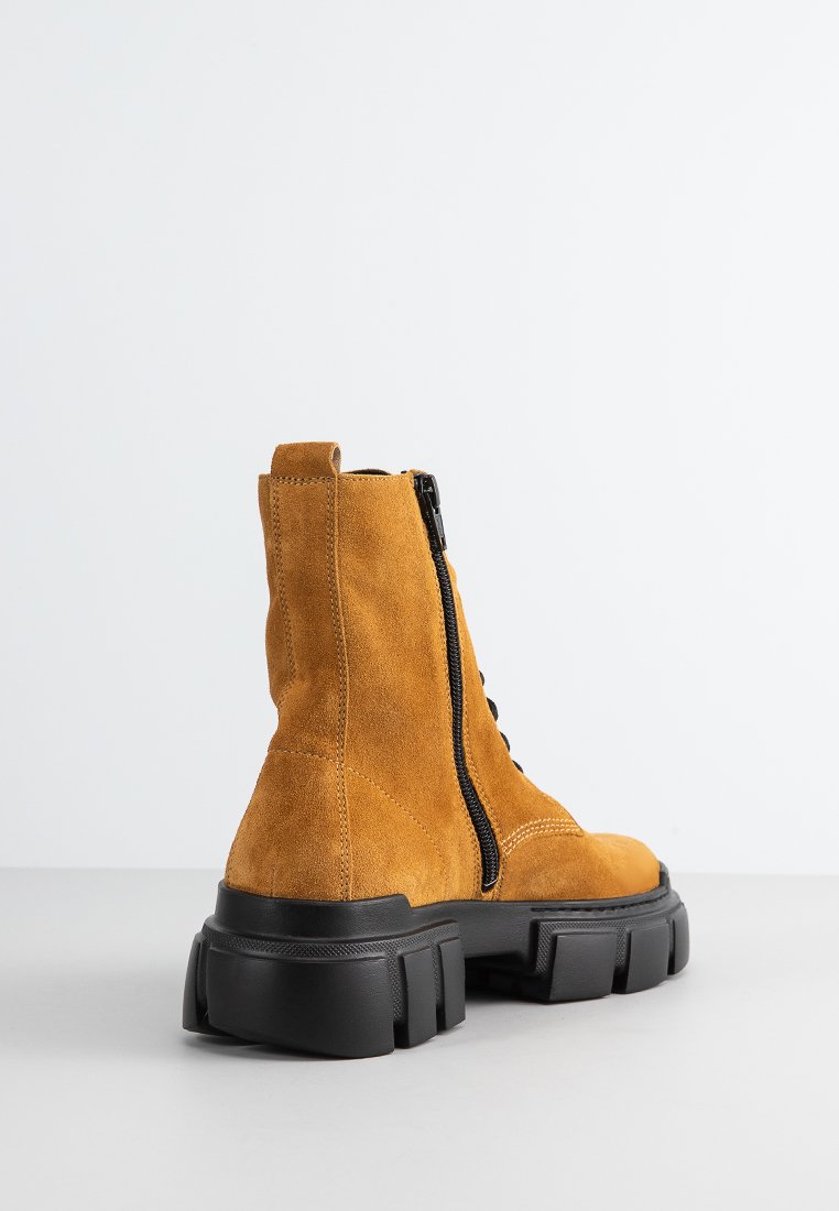 Botas de ante en amarillo mostaza con talón acolchado, suela de goma negra, cremallera lateral y detalles de cordones en la parte delantera.