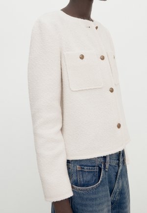 Person wearing a white textured cropped jacket with brown buttons and front flap pockets, paired with blue denim jeans.