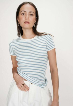 Femme portant un haut à manches courtes rayé bleu et blanc, pantalon blanc taille haute, boucles d'oreilles en or et bagues, debout devant un fond uni.