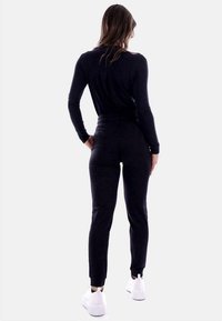 Zwarte longsleeve bodysuit met een aansluitend ontwerp, gecombineerd met een bijpassende zwarte joggingsbroek en witte sneakers, gezien vanaf de achterkant.