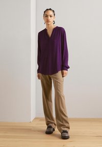Purple long-sleeve blouse with a V-neck, paired with light beige wide-leg trousers and dark shoes, set against a neutral background.