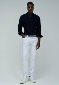 Camisa de botones azul marino, jeans ajustados blancos y zapatillas blancas. El conjunto incluye un cinturón estampado y texturas suaves.