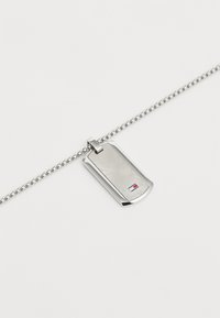 Pendentif en forme de plaque militaire en argent avec une surface texturée et un petit logo. Attaché à une chaîne à perles pour un look élégant.