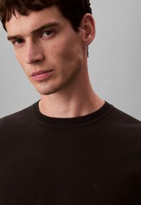 Mladý muž s krátkými hnědými vlasy, oblečený v černém svetr s kulatým výstřihem a jemným logem "cK" na hrudi, na šedém pozadí.