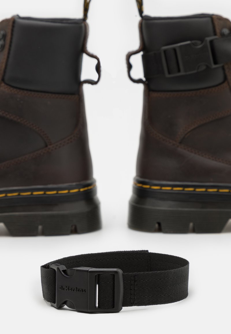 Donkerbruine leren laarzen met zwarte accenten, gele stiksels, een robuuste zool en twee voorste gespen; vergezeld van een zwarte band met een plastic gesp.