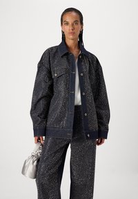 Chaqueta de mezclilla oscura con patrón de tachuelas plateadas, corte oversized, gran cuello y cierre de botones. Combinada con pantalones a juego y un bolso plateado.
