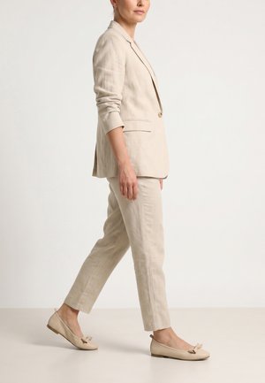 Kvinde går i beige linnedragt med skræddersyet blazer, korte bukser og matchende ballerinaer med sløjfedetalje.