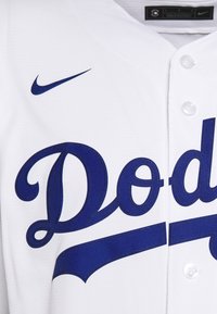 Vit basebolltröja med blå "Dodgers"-bokstäver, blå Nike-logga, knäppning framtill och textilstruktur.