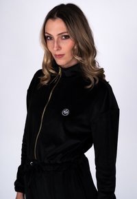 Vrouw met golvend blond haar, gekleed in een zwart shirt met rits en een klein wit cirkelvormig embleem op de borst, staand tegen een effen witte achtergrond.