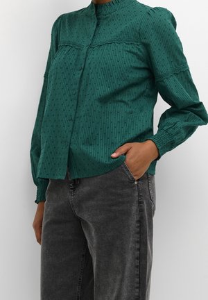 Vrouw die een donkergroene, gestructureerde blouse met knopen draagt, voorzien van kanten rand en geplooide manchetten, gecombineerd met vervaagde zwarte spijkerbroek, hand in zak.