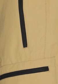 Nahaufnahme eines beigen Stoffs mit zwei schwarzen Reißverschlüssen, die vertikal und horizontal verlaufen, mit detaillierter Nahtführung rund um die Reißverschlüsse.