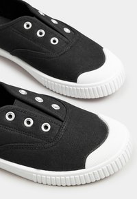 Scarpe slip-on nere in tela con punta e suola in gomma bianca, dotate di cinque occhielli metallici per lacci su una tomaia nera testurizzata.