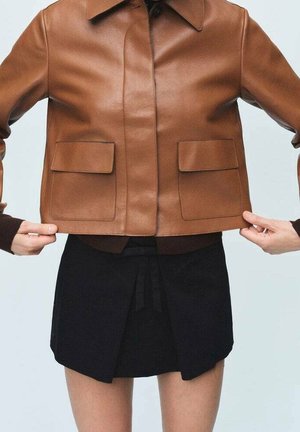 Persona con una chaqueta de cuero marrón con bolsillos con solapa, sujetando el dobladillo, y una mini falda negra con pliegues en la parte delantera.