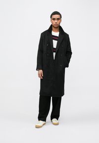 Sort dobbeltradet frakke med slagkrave, kombineret med en strikket, stribet sweater, sorte bukser og hvide sneakers med beige såler.