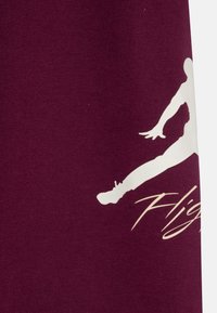 Bordeauxfarbener Stoff mit einer großen weißen Silhouette einer springenden Figur und dem Text "Flug" in einer geschwungenen Schriftart. Glatte Textur, sportliches Design.